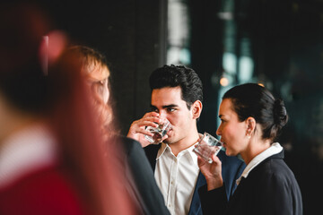 Professionals in formal attire network at an indoor event, sipping water and conversing in a dimly lit lounge.