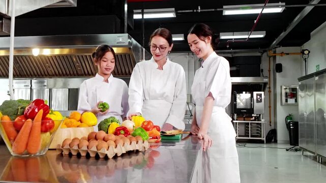 Chef instructor teaching two young culinary students in a commercial kitchen during a professional cooking class with fresh vegetables. Ideal for chef training, gastronomy, and food education themes.