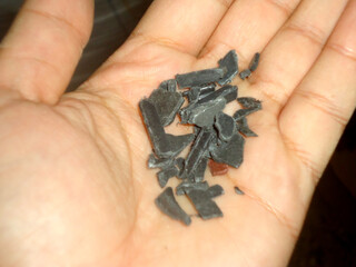 A handful of dark, irregularly shaped, crushed plastic fragments, or flakes, held in an open palm. Represents the concept of plastic recycling, waste management, or sustainability efforts.