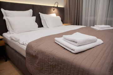 Neatly made hotel bed with folded white towels on a beige quilted blanket in a cozy modern bedroom. Warm lighting and soft textures create a calm, welcoming atmosphere.