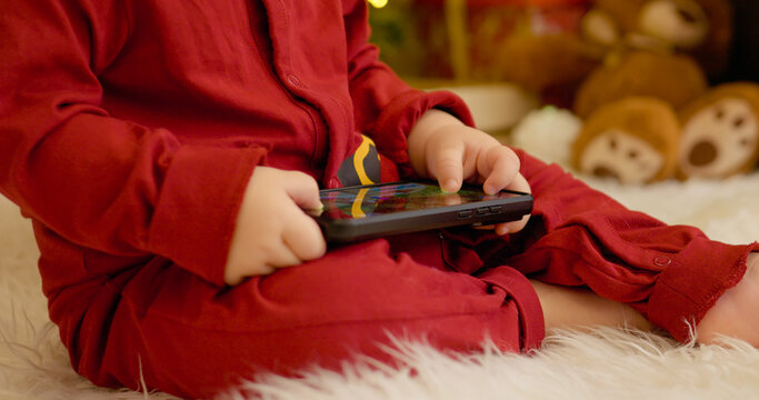 A young boy sits comfortably on the floor, focused on his smartphone as he enjoys the festive atmosphere created by the Christmas tree and colorful decorations.