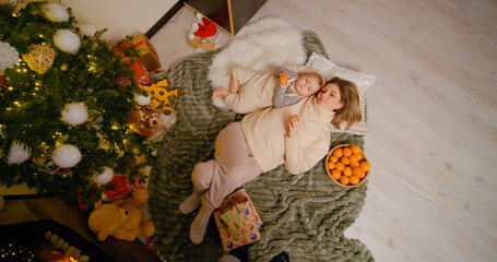 A little boy snuggles with his mother on a blanket next to a beautifully decorated Christmas tree,...