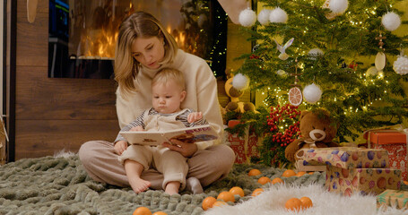 A young mother and her toddler sit comfortably on a blanket, sharing stories from a book while...