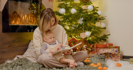 Under the glow of the Christmas tree, a young mother and her little boy share a warm moment as they read stories together, surrounded by festive decorations and toys.
