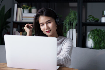 Naklejka premium Asian woman sits on sofa wearing headphone watching movie on laptop in living room