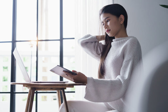 Asian woman sits on sofa using digitalized pen writing on tablet with laptop on table in living room