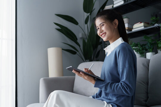Asian woman sits on sofa using digitalized pen write on tablet in living room