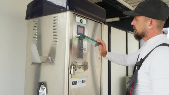 Man in overalls at self-service car wash inserting token into payment machine and pressing start button to activate automatic vehicle cleaning cycle