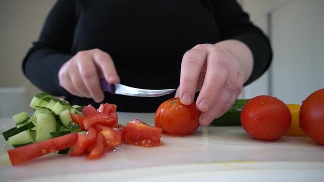 Slicing vegetables on a board. Hands cutting tomatoes with a knife, next to cucumber slices, home cooking, close-up. Video without a face.