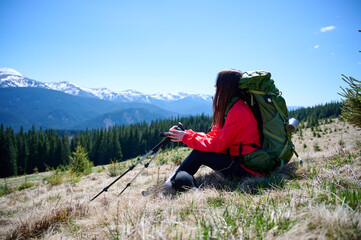 Female hiker resting on grassy mountain slope with trekking poles. Woman in red jacket enjoying scenic view of snowy peaks and sunny weather. Outdoor travel and adventure lifestyle.