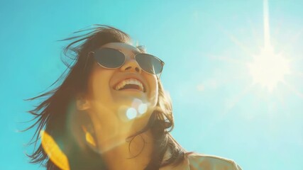 A joyful young lady wearing sunglasses and a sun dress smiling brightly on a clear blue day. She is facing the camera with her hair blowing in the wind.