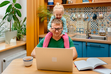 A young father is working on a laptop at home in the kitchen, playful daughter little child is...
