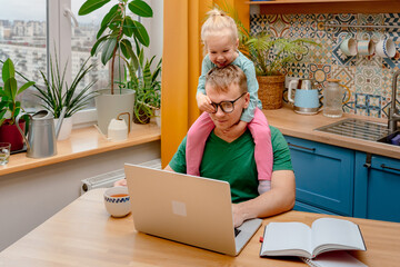 A young father is working on a laptop at home in the kitchen, playful daughter little child is...