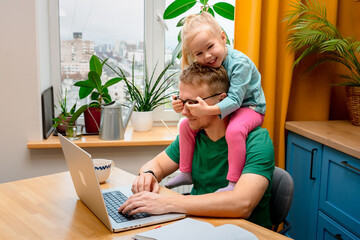 A young father is working on a laptop at home in the kitchen, playful daughter little child is...