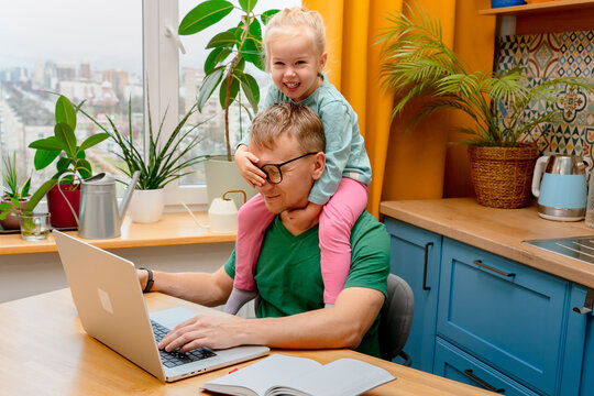 A young father is working on a laptop at home in the kitchen, playful daughter little child is sitting on his neck, preventing him from working