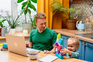 Father works remotely in the kitchen on a laptop, daughter little child plays with a construction...