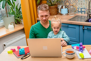Father works remotely in the kitchen on a laptop, daughter little child distracts attention...