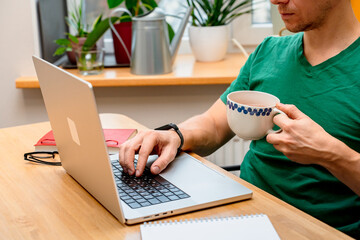 A young man works from home in the kitchen using a laptop. Freelancer working from home