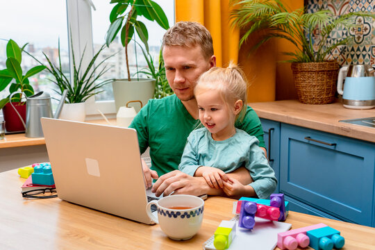 Father works remotely in the kitchen on a laptop, daughter little child distracts attention disturbs parent from work on laptop