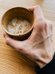 First-person view: close up of hand gently holding disposable cup of cappuccino, featuring delicate latte art design, resting on warm toned wooden table in coffee shop setting. Hand gripps cappuccino