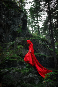woman wearing a long red cloak gracefully walks down a rocky path surrounded by green moss and trees in a dimly lit forest. atmosphere is serene and mysterious