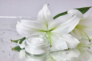 small jar of skincare cream is placed next to a fresh white lily flower on a shiny surface,...