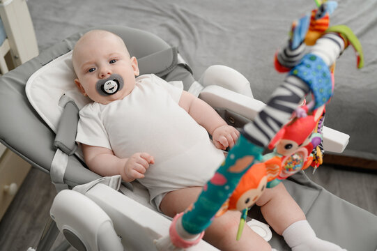 Peacefully playing baby lies in comfortable rocking chair with toys for entertainment, sucking on pacifier. Infant enjoys quiet play surrounded by soothing rattles, feeling safe.
