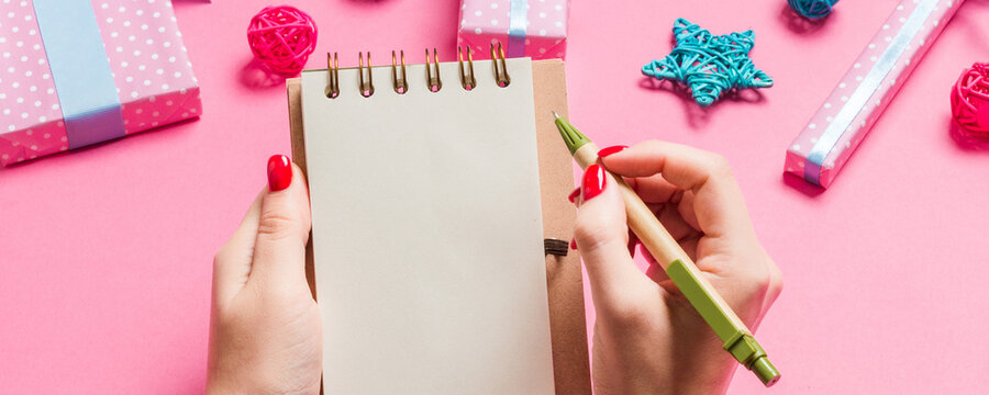 Top view of female hand making some notes in noteebok on pink background. New Year decorations and toys. Christmas time concept