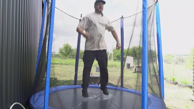 An adult man jumping on a trampoline. He is wearing a T-shirt and black pants. The trampoline is outside. A country house. Having fun. Relaxing. Sports. Daylight.