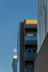 Full moon appears in blue sky over buildings catching last warm light of day as they disappear into shadows of surrounding buildings during sunset.