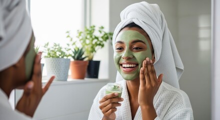 A happy woman in a white bathrobe and towel applies a green facial mask, smiling at her reflection. The bright, natural light creates a fresh, relaxed atmosphere.