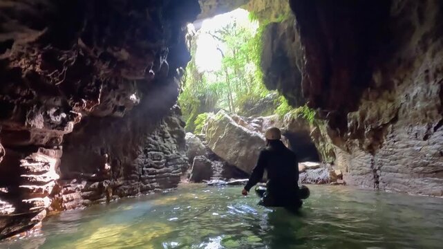 A man emerges from the exit of a rugged cave in Waitomo, New Zealand. Stunning rock formations, stalagmites, and natural scenery highlight the dramatic underground landscape.