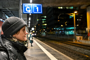 Woman in a black winter jacket and beanie waits on a night platform in Zurich