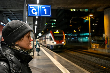 Woman in a black winter jacket and beanie waits on a night platform in Zurich. A train stands beside her under bright station lights in an urban setting