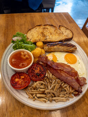 A hearty breakfast spread with crispy bacon, fried eggs, grilled sausage, baked beans, sautéed mushrooms, charred tomatoes, golden hash browns, and toasted sourdough bread on a rustic wooden table.