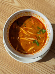 Vibrant and aromatic Tom Yum soup, a classic Thai dish, served in a white bowl on a sunlit wooden table. This spicy and sour broth is packed with fresh ingredients, perfect for an authentic culinary