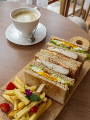 A delectable club sandwich with fried egg, chicken, and fresh vegetables, served with crispy french fries, cherry tomatoes, and a hot cup of coffee on a wooden table. Perfect for a morning or