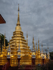 Naklejka premium Golden Buddhist chedi shimmering at dusk, surrounded by ornate spires and glowing lights. Serene evening view of Wat Phra Singh Woramahawihan in Chiang Mai, Thailand, a revered spiritual landmark.