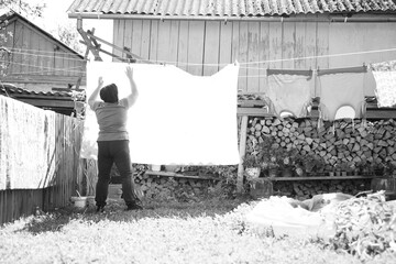 Monochrome shot captures a person hanging laundry in a backyard, showcasing domestic tasks and simple rural living in black and white.
