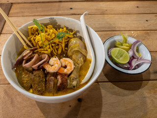 Vibrant bowl of authentic Northern Thai Khao Soi, a rich curry noodle soup with chicken, shrimp, squid, and crispy toppings, served on a rustic wooden table with fresh condiments.