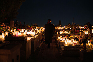 Night scene of a cemetery lit by hundreds of candles with a person walking along the path. Warm lights, flowers and glowing lanterns create a peaceful and atmospheric All Saints Day setting in Biały K