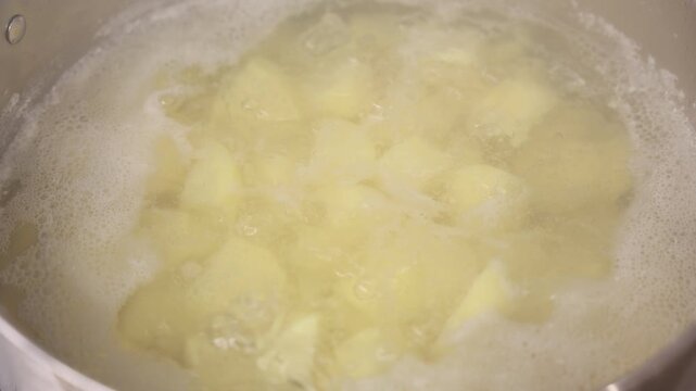 Close-up view of peeled, chopped potatoes boiling in a pot of water, surrounded by foam and bubbles during the cooking process