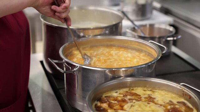 A cook stirs a large pot of boiling vegetable soup with shredded carrots and cabbage in a commercial kitchen, surrounded by other simmering pots