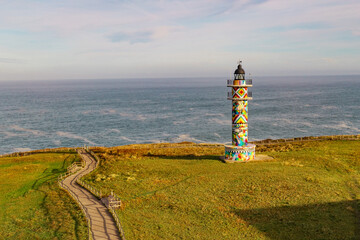 Colorful Ajo Lighthouse captured by drone, standing on a green cliff with a vast ocean backdrop. Coastal landscape of Faro de Ajo in Cantabria, Spain