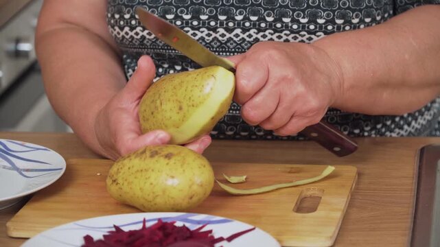 Peeling potato with knife for cooking Ukrainian borscht. Removing skin from fresh tuber for preparing traditional soup. Cleaning raw potato for making borscht recipe. Preparing starchy vegetable for