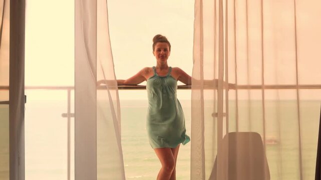 Vacation Woman Tourist Stands on the Balcony of a Resort Room with Beautiful Ocean View at the Yellow Sunset, Everning Window View, Slow Motion. Vacation, Travel, Summer Holidays, Tourism.