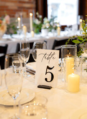 Elegant wedding reception table setup with “Table 5” sign, candles, and glassware on a white tablecloth. Romantic fine dining atmosphere with warm candlelight and floral decor in the background