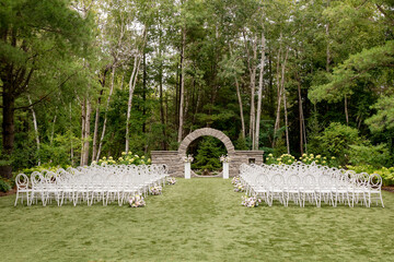 Elegant outdoor wedding ceremony setup framed by a lush forest. White chairs line the aisle leading...