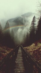 A wooden bridge leads through a misty forest to the mountains. A rainbow arches across the sky, adding a touch of magic to the scene.