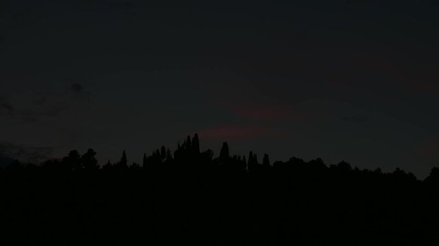 Pink and grey clouds glide over the sky above a shadowy forest outline as the sun sets. Gradually, the sky transitions from evening light to complete darkness